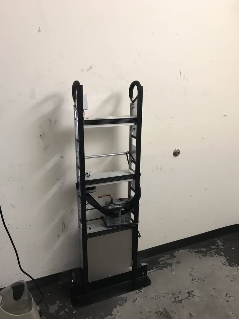 A black dolly with straps stands against a wall in a room with scuffed floors, likely used for transporting indoor HVAC equipment.