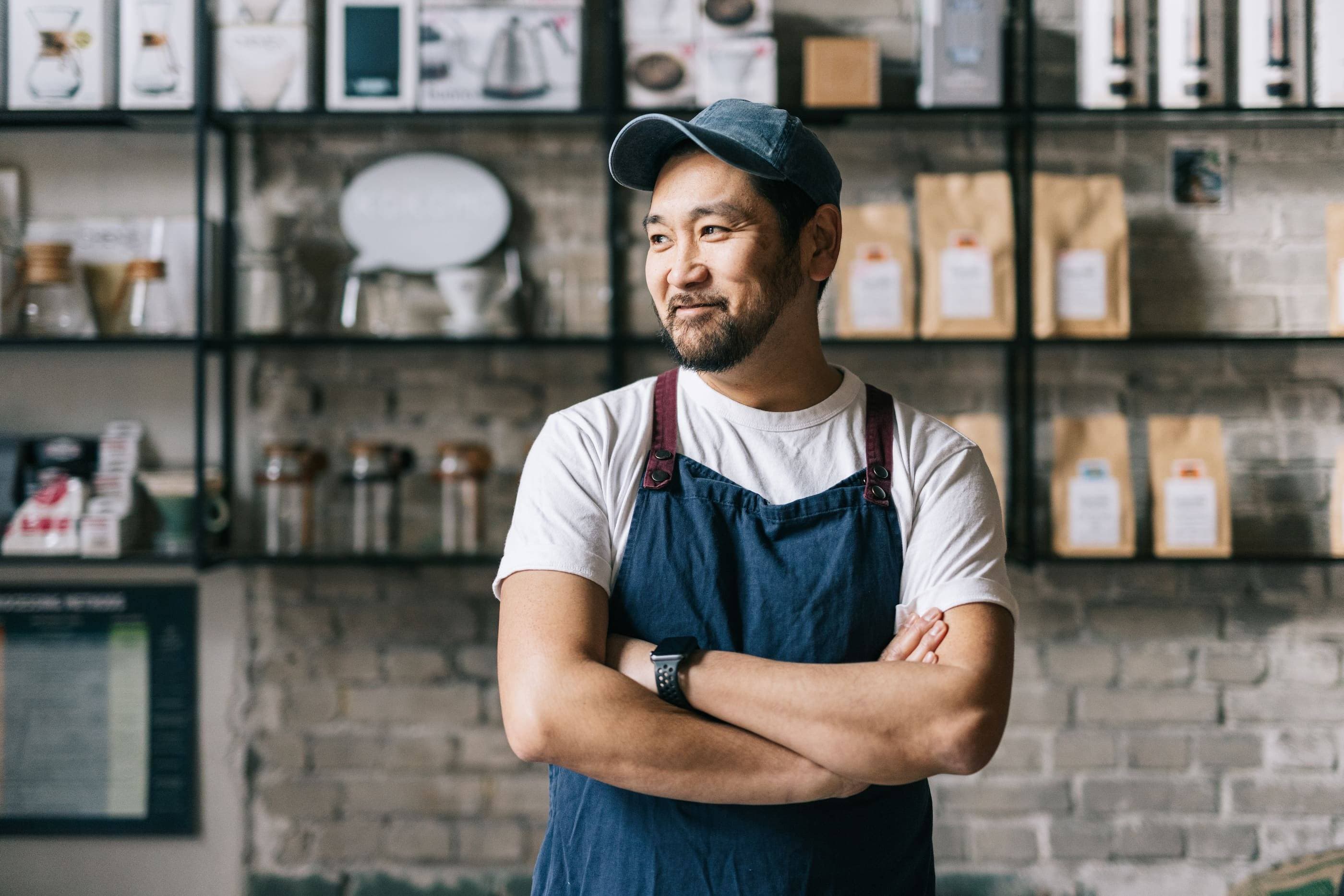 A young asian man in a hat and apron standing in a coffee shop.