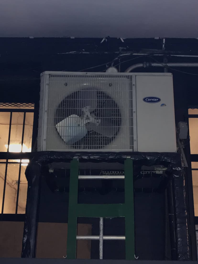 An air conditioning unit, specifically one of the modern mini-split systems, is mounted on a green metal stand with a window and pipes elegantly in the background.