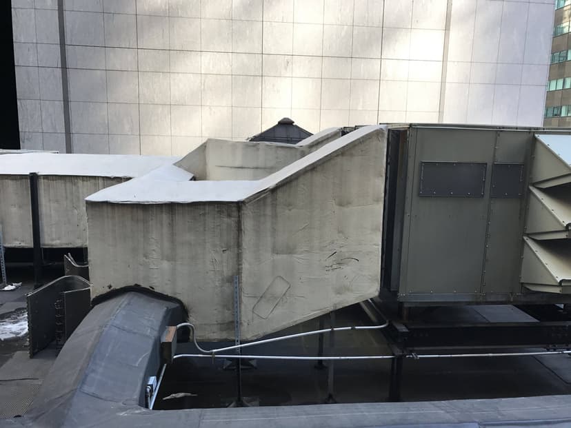 Rooftop ventilation system with large ducts and an industrial unit against a concrete building backdrop, featuring commercial HVAC outdoor units.