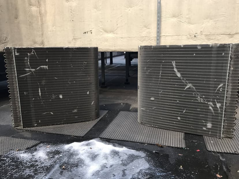 Two large curved metal air conditioning coils on the ground, next to a building, resemble commercial HVAC outdoor units. Snow and ice are around the coils.