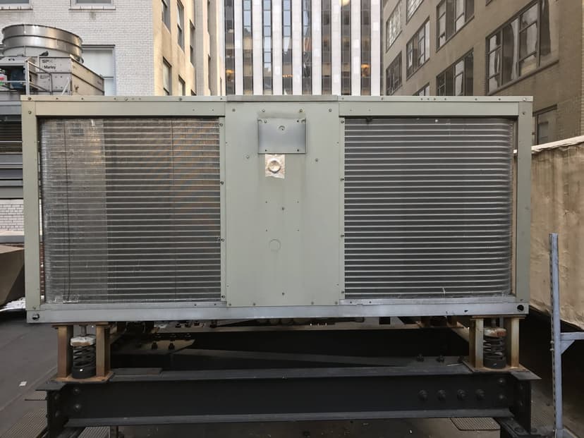 A large commercial HVAC outdoor unit is located on a building rooftop, surrounded by tall buildings.