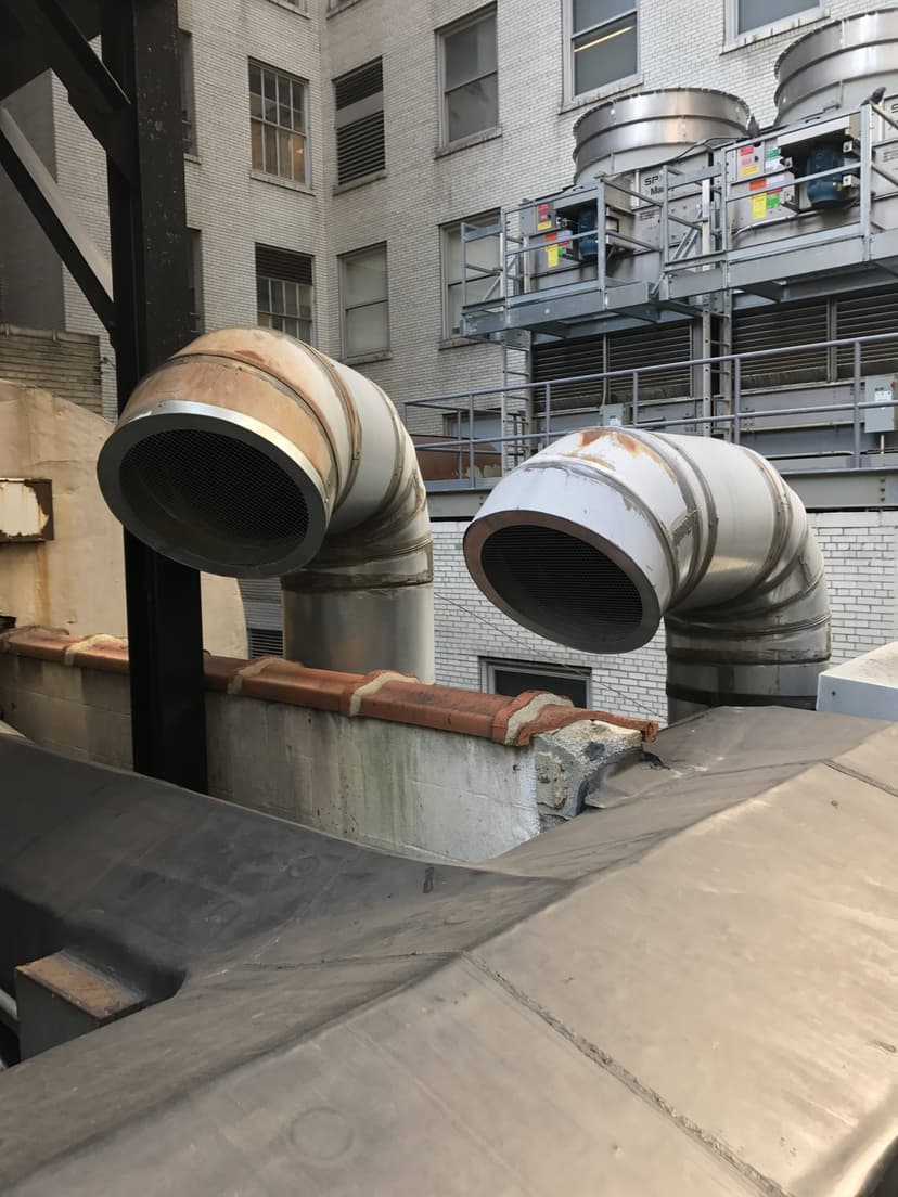 Large industrial ventilation ducts and commercial HVAC outdoor units on the rooftop of a commercial building against an urban background.