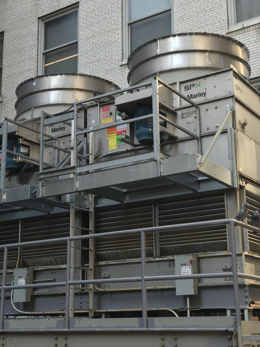 Two industrial cooling towers against a brick building wall, marked with 'SPX Marley' and warning labels, sit alongside commercial HVAC outdoor units.