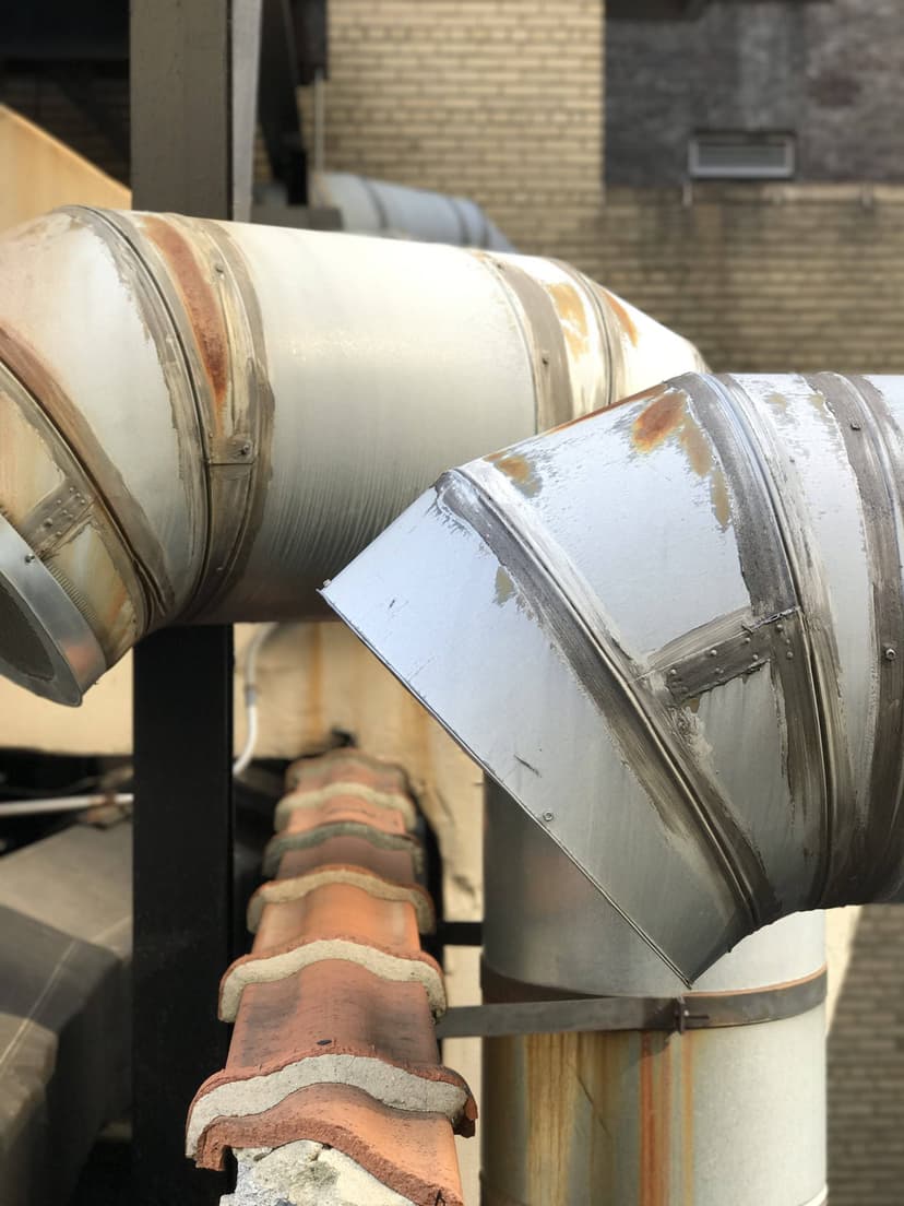 Close-up of large, rusted, metallic HVAC ducts and commercial HVAC outdoor units against a brick wall on a rooftop.