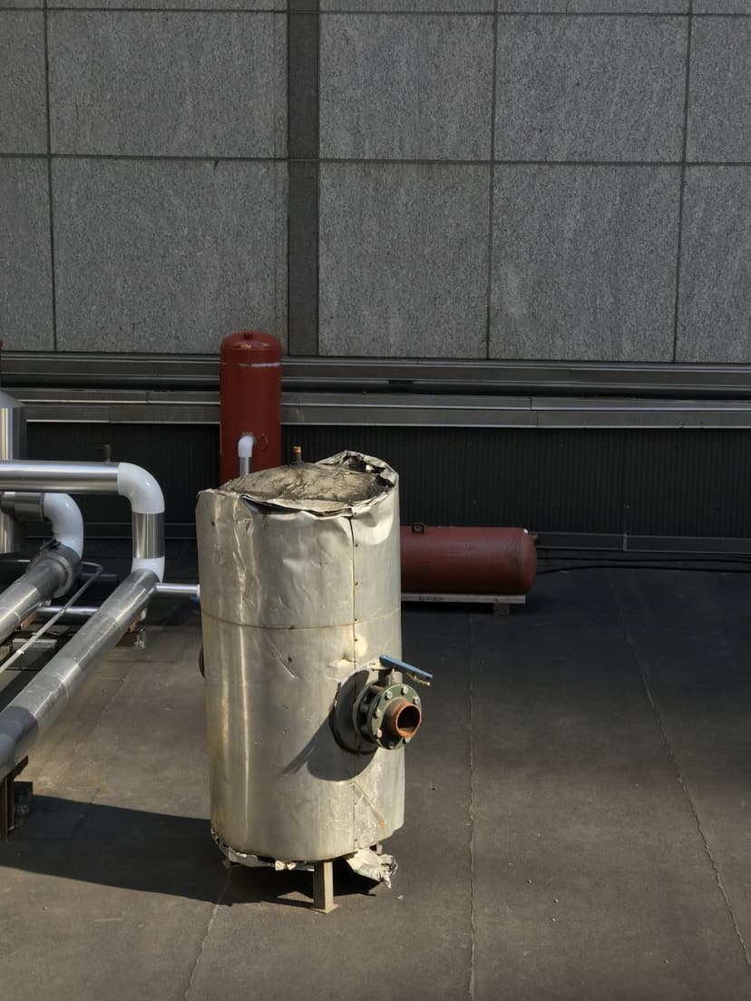 A damaged metal cylinder with pipes is on a rooftop near a wall, alongside commercial HVAC outdoor units, with a cylindrical red object in the background.