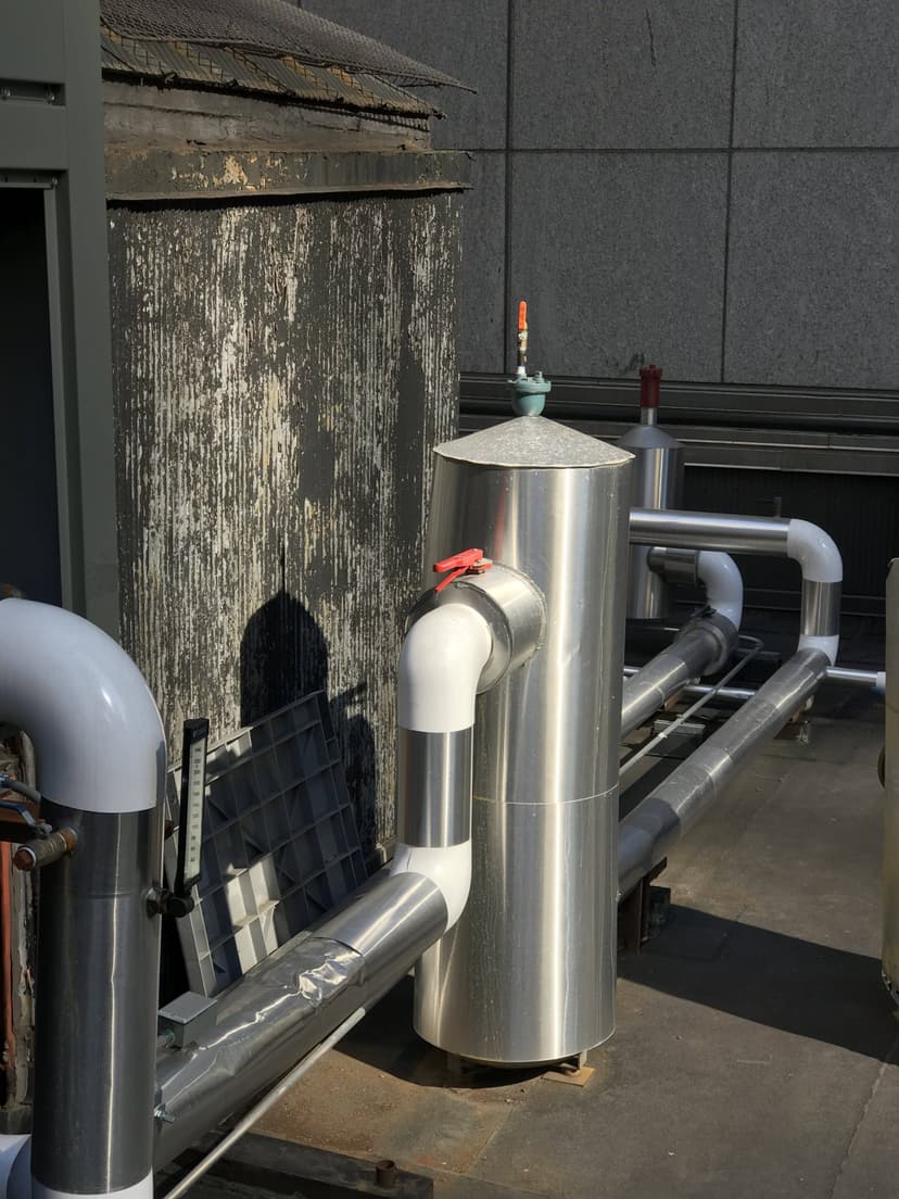 An industrial setup with insulated metal pipes and a valve on a rooftop, alongside commercial HVAC outdoor units, placed near a weathered building wall.