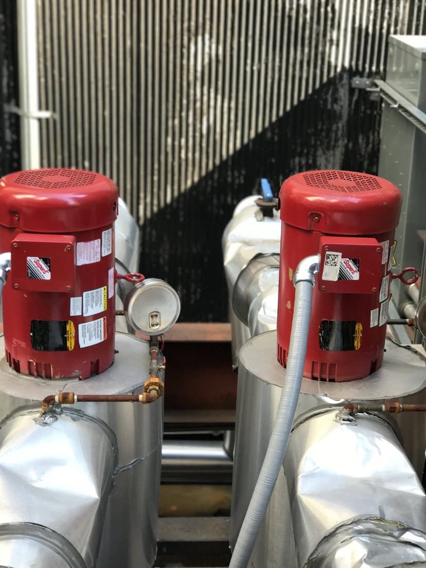 Two red industrial pumps connected by pipes, with gauges and labels, set in front of a corrugated metal background resembling commercial HVAC outdoor units.