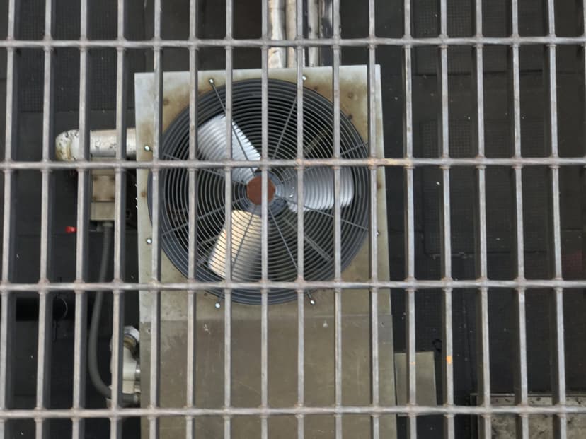 A large industrial fan viewed through a metal grate, with pipes and machinery partially visible in the background, reminiscent of commercial HVAC outdoor units.