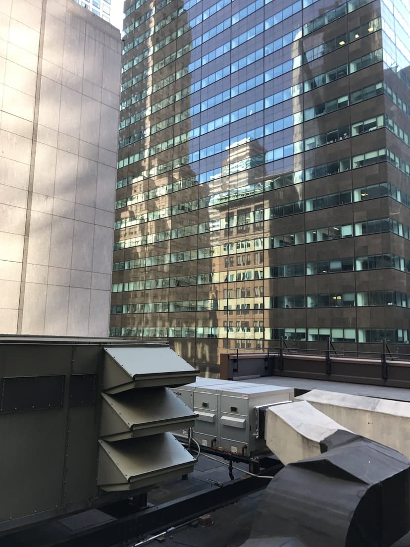 Cityscape with a reflective glass building, adjacent concrete structures, and rooftop ventilation systems alongside commercial HVAC outdoor units in the foreground.