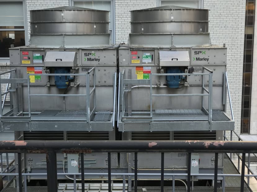 Two commercial HVAC outdoor units, adorned with various colored labels and a SPX Marley logo, rest on a metal platform against a brick wall.