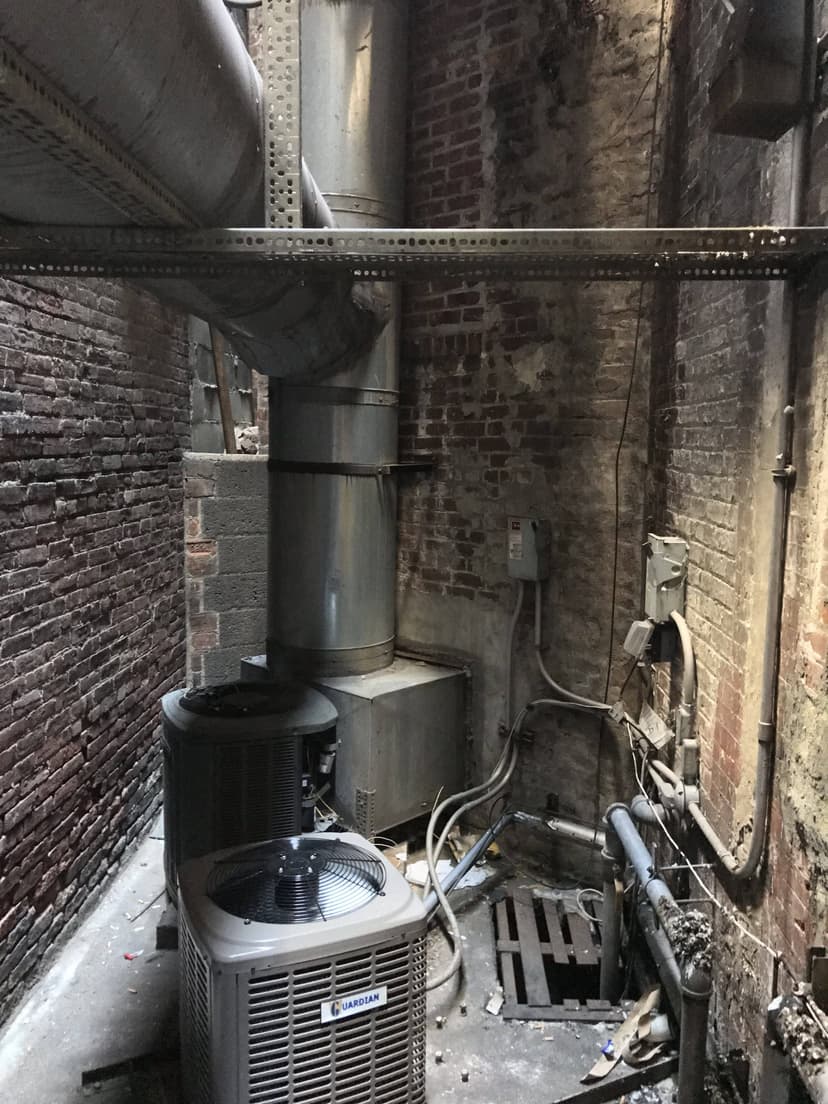 An industrial alley with commercial HVAC outdoor units and exposed ductwork against brick walls.