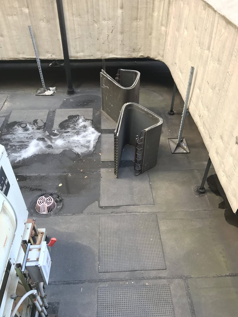 Concrete ledge overlooking an industrial area with metal barriers, grates, pipes, and a drain, dotted with commercial HVAC outdoor units nearby.