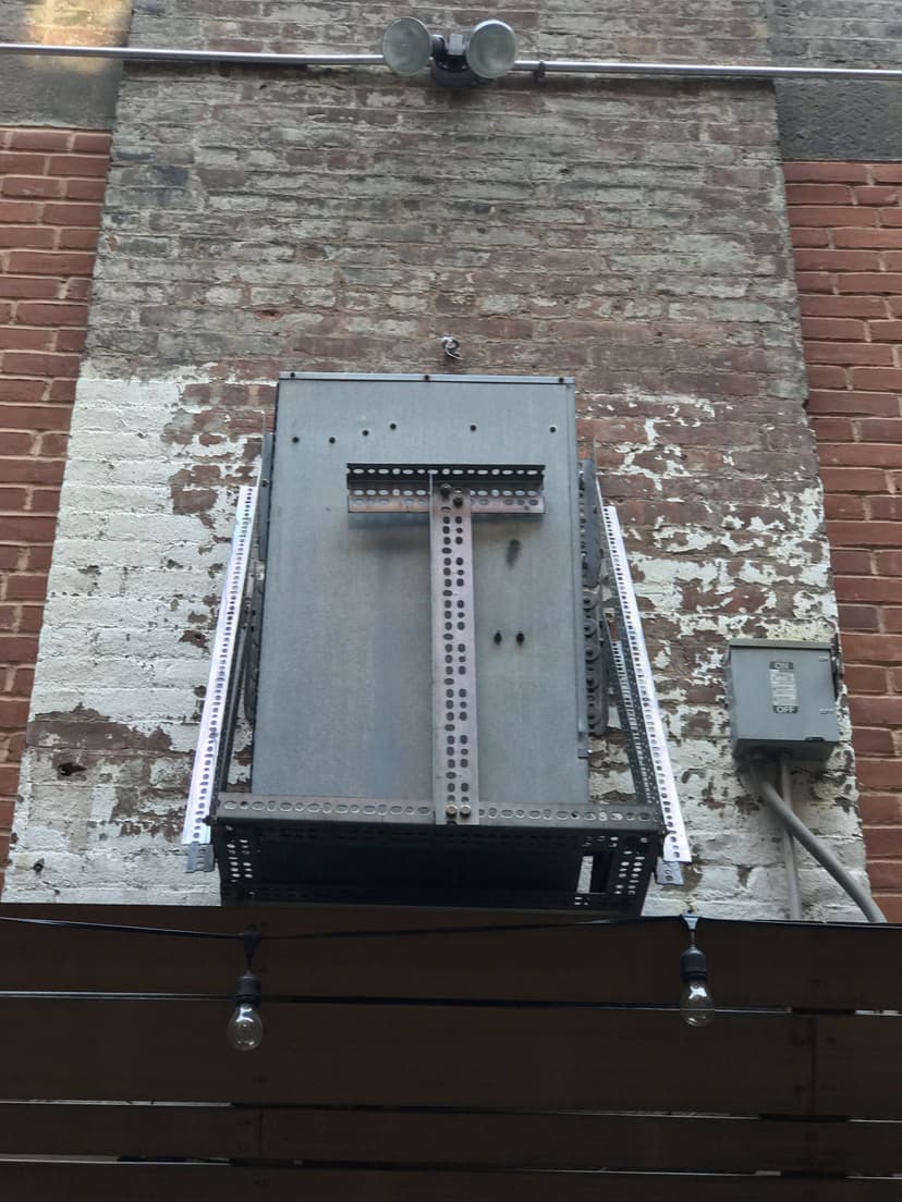 A metal box and conduit attached to a brick wall, with two light bulbs on either side below it, harmoniously coexist alongside commercial HVAC outdoor units, ensuring optimal functionality.