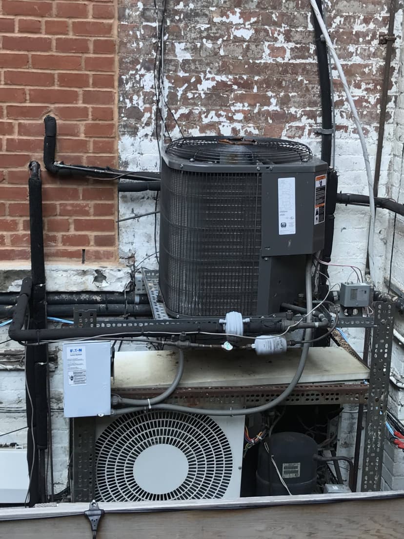 A commercial HVAC outdoor unit is mounted on a metal stand against a brick and painted wall, complete with electrical and cooling components.