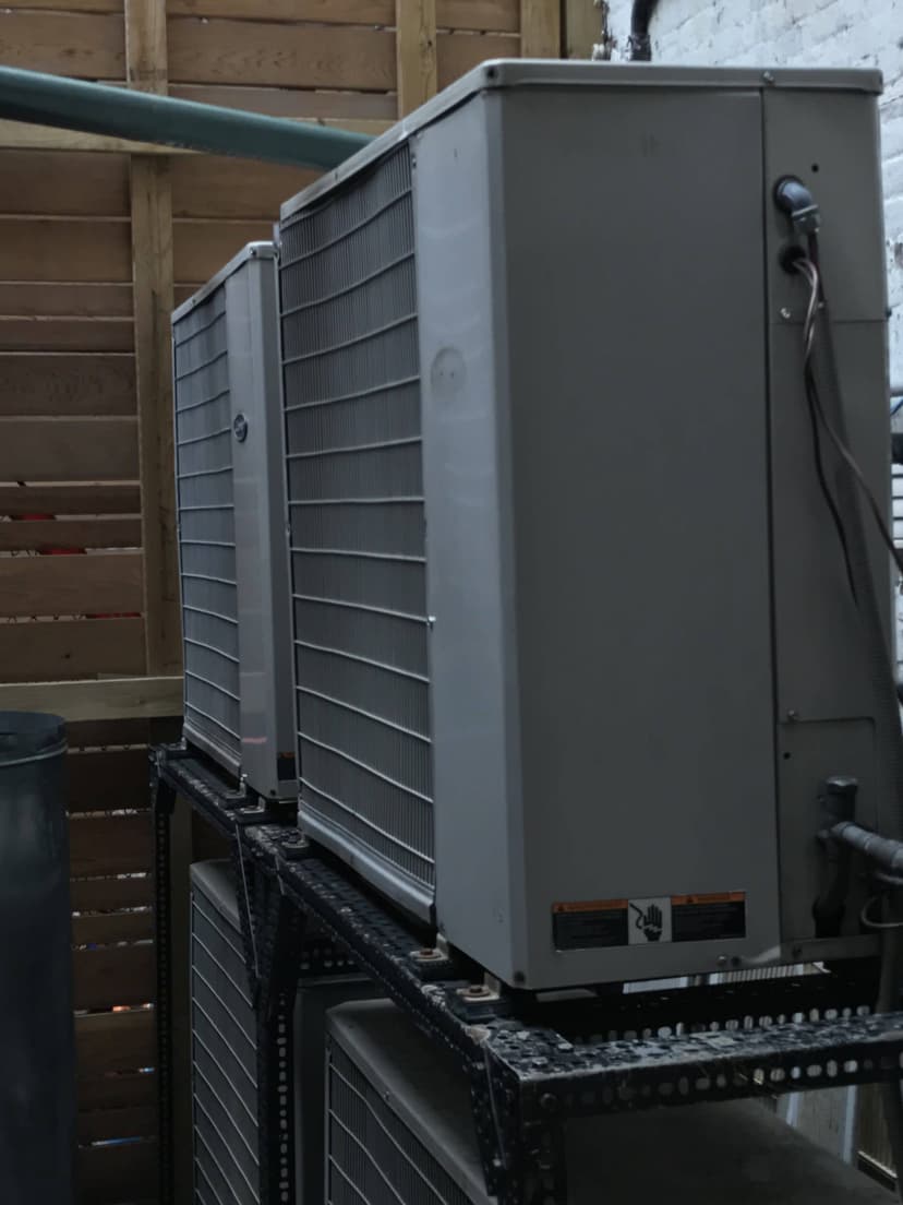 Two large commercial HVAC outdoor units are mounted on a metal stand, positioned against a wooden fence.