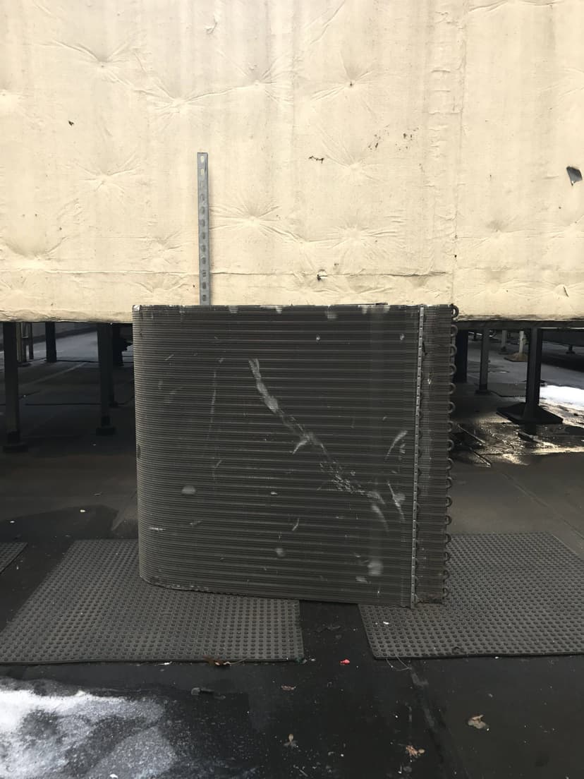 A large industrial cooling coil with visible wear and dirt stands in front of an insulated structure, reminiscent of commercial HVAC outdoor units.