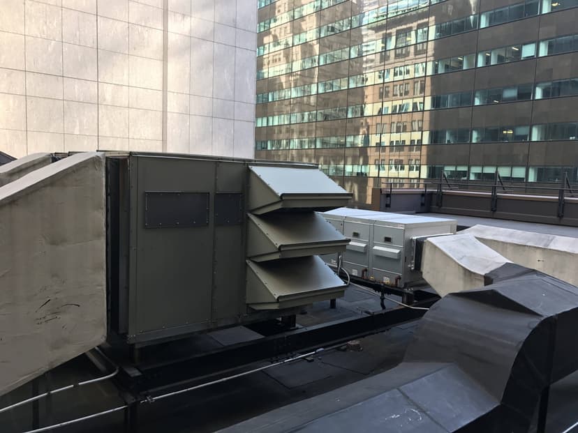 Large packaged unit HVAC systems on a rooftop are surrounded by tall office buildings with glass windows in a bustling city setting.