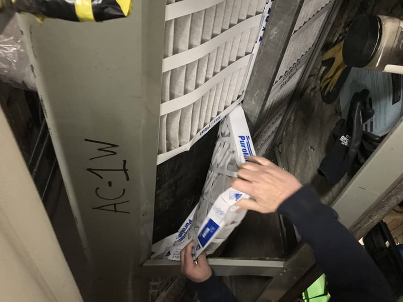 A person performing preventative maintenance for HVAC is replacing an air filter in a unit labeled 'AC-1W,' their hands carefully holding the new filter.