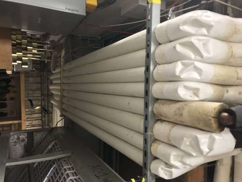 A stack of large, rolled, and wrapped white tubes stored on metal shelves in an industrial setting, crucial for preventative maintenance for HVAC systems.