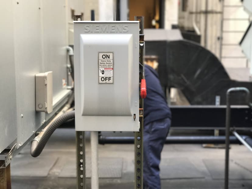 A person in blue work attire operates near a Siemens electrical switch box with an 'ON/OFF' label in an industrial setting, with a rooftop HVAC system visible in the background.