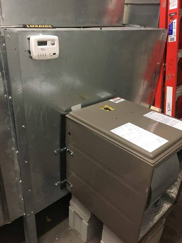 An HVAC unit with a LUXAIRE thermostat attached to a metal surface features steam coils and a gray control box on concrete blocks below.