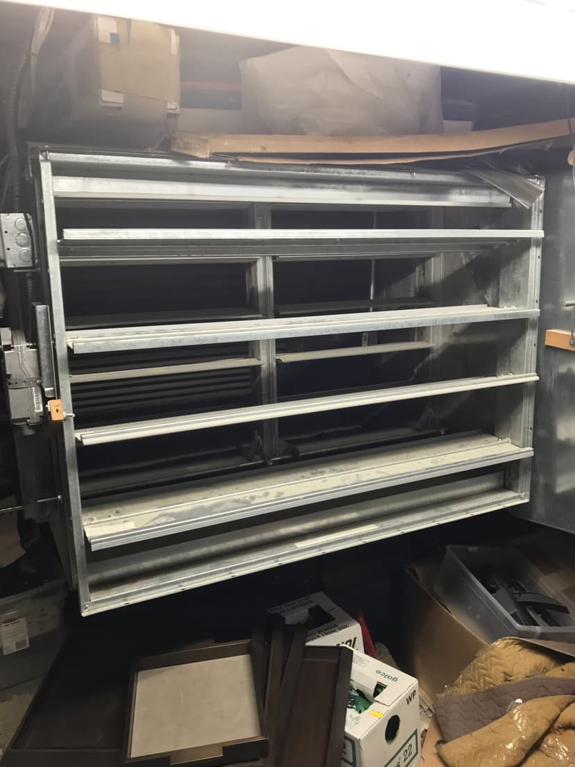 Large metallic air duct with multiple horizontal aluminum louvers inside a cluttered room, boxes and papers nearby.