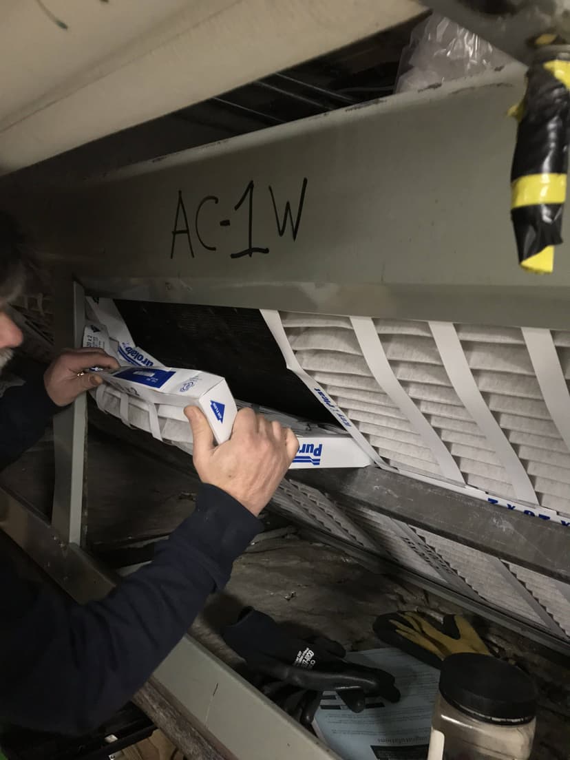 A person changing HVAC filters labeled AC-1W under a metal structure.
